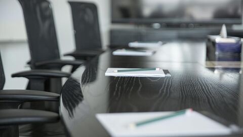 Bildausschnitt eines runden Konferenztisches mit Stühlen. Auf dem Tisch liegen Papier und Stifte.